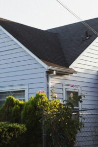 A homeowner examines roof shingles and clear gutters from the ground beside a well-maintained house.